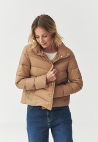 Beige pufferjack met hoge kraag, drukknoopsluiting en horizontaal gestikt patroon, gedragen over een lichte top en gecombineerd met blauwe jeans.