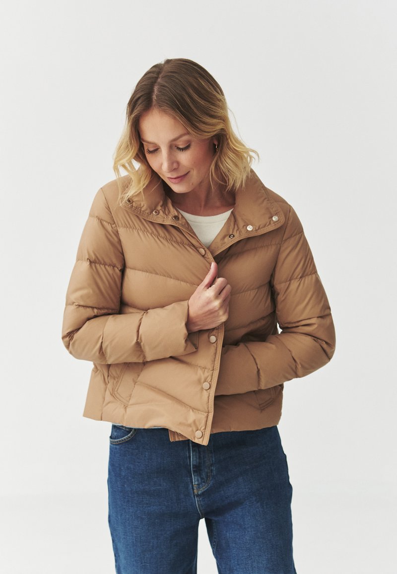 Beige pufferjack met hoge kraag, drukknoopsluiting en horizontaal gestikt patroon, gedragen over een lichte top en gecombineerd met blauwe jeans.