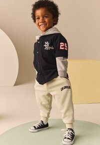 Veste noire à manches courtes avec capuche grise, broderie blanche et accents rouges, associée à un pantalon de jogging crème et des baskets noires.