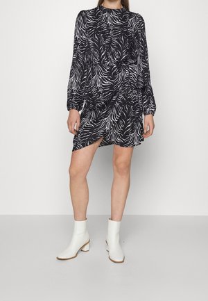 Vestido negro con estampado de cebra blanco, mangas largas, cuello alto, diseño drapeado y abertura lateral, combinado con botas blancas de tobillo.