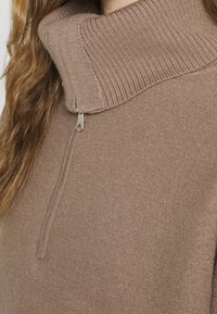 Brązowy dzianinowy sweter z wysokim kołnierzem, z zamkiem błyskawicznym. Miękka tekstura i prążkowane wykończenia na kołnierzu i dole.