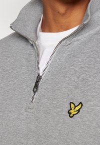 Felpa grigia con zip e colletto alto, con un logo ricamato di un uccello giallo e nero sul petto a sinistra e una zip argentata.