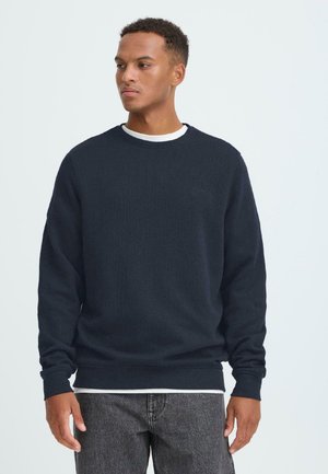 Un sweat-shirt bleu marine à col rond en tissu doux, avec des manches longues et des poignets côtelés, superposé sur une chemise blanche.