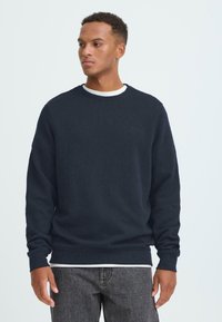 Sudadera de cuello redondo azul marino hecha de tejido suave, con mangas largas y puños acanalados, superpuesta sobre una camisa blanca.