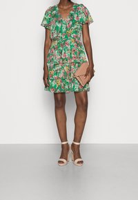 Bloemige groene jurk met korte mouwen, V-hals, gelaagd ontwerp en levendige patronen. Gecombineerd met nude hakken en een bijpassende clutch.