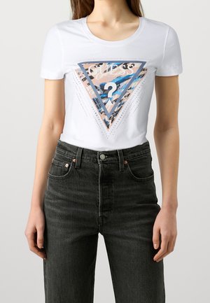 Femme portant un t-shirt blanc Guess avec un logo triangulaire coloré et un jean noir taille haute, debout sur un fond uni.