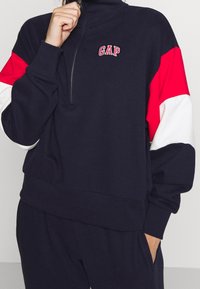 Felpa con zip di colore blu navy con maniche rosse e bianche. Presenta il logo "GAP" sul petto e polsini e orlo a costine. Tessuto morbido e strutturato.