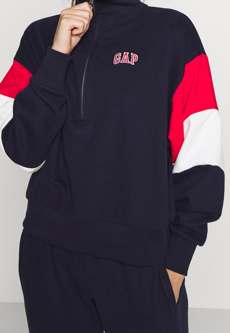 Felpa con zip di colore blu navy con maniche rosse e bianche. Presenta il logo "GAP" sul petto e polsini e orlo a costine. Tessuto morbido e strutturato.