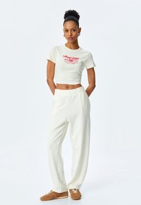 Crème cropped t-shirt met rode tekst, gecombineerd met witte relaxte sweatpants. Hoogtepunten zijn het eenvoudige ontwerp en de zachte textuur.