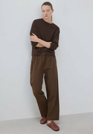 Femme à la peau claire et aux cheveux bruns attachés, debout les bras croisés, portant un haut à manches longues marron foncé, un pantalon large marron et des chaussures marron sans lacets.