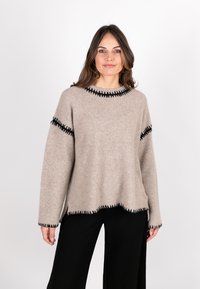 Beiger Wollpullover mit Rundhalsausschnitt, schwarzen Nähten entlang der Kanten und lockerem Schnitt. Kombiniert mit schwarzen weiten Hosen.