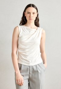 Hladký bezrukávový top z jemnej, svetlo béžovej látky s okrúhlym výstrihom. V kombinácii s svetlošedými pruhovanými nohavicami s voľným strihom.