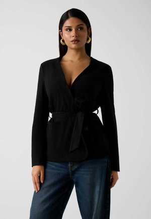 Blusa negra envolvente con mangas largas, cintura ceñida, escote en V y dos bolsillos delanteros. Combinada con vaqueros azules holgados. Textura de tela suave.