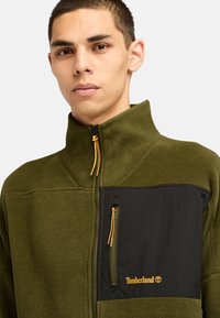 Giacca in fleece verde oliva con una tasca sul petto nera. Caratterizzata da collettore alto, chiusura a zip e cerniere gialle. Logo Timberland in oro.