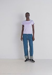 T-shirt violet clair avec le texte "Pepe", associé à un jean bleu retroussé et des chaussures de sport noires, présenté sur un fond uni.