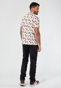 Camicia a maniche corte color crema con motivo di insetti neri e rossi, abbinata a pantaloni neri e sneakers con accenti rossi.