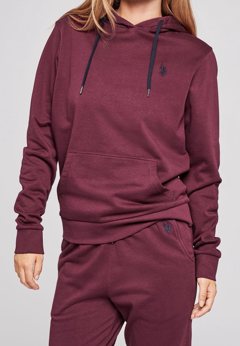 Sweat à capuche bordeaux en tissu doux avec poche kangourou, capuche à cordon de serrage, et logo sur la poitrine, accompagné d'un pantalon de survêtement assorti.