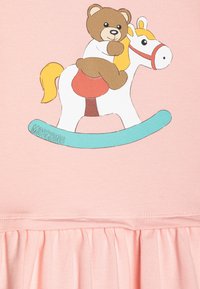 Vaaleanpunainen mekko, jossa on karhu keinutuolissa -grafiikka. Suunnittelussa on värikkäitä yksityiskohtia, röyhelöhelma ja sileä kangaslaatu.