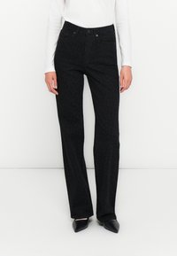 Woman wearing high-waist black pants with subtle leopard print, white long-sleeve top, and black pointed-toe shoes against plain background.