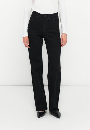 Woman wearing high-waist black pants with subtle leopard print, white long-sleeve top, and black pointed-toe shoes against plain background.