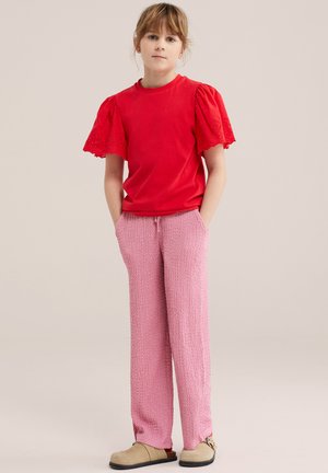 Młoda osoba stojąca z rękami w kieszeniach, ubrana w czerwony top z bufkami, różowe teksturowane spodnie oraz beżowe buty wsuwane.