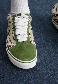 Zapatilla con una puntera de ante verde, cordones blancos y un panel lateral texturizado en marrón y blanco, sobre una alfombra azul oscura.