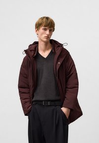 Giacca a vento bordeaux con cappuccio, caratterizzata da una texture lucida e chiusura con zip, indossata sopra un maglione scuro a V e pantaloni neri con cintura.