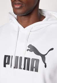 Hvid sweatshirt med kengurulomme, justerbar hætte og et fremtrædende sort logo med teksten "PUMA" samt et springende sort katteemblem.