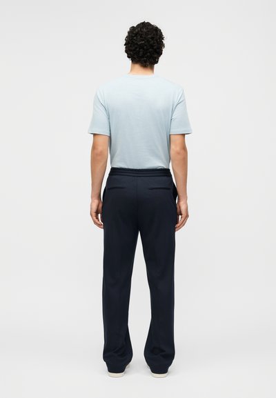 Άντρας με σγουρά μαλλιά στέκεται πλάτη, φοράει ανοιχτό μπλε κοντομάνικο πουκάμισο, σκούρο μπλε παντελόνι και λευκά παπούτσια σε μονόχρωμο φόντο.