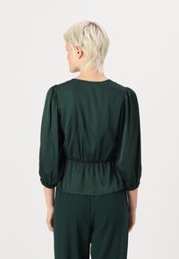 Mørkegrønn satengbluse med pufferm og elastisk midje; glatt tekstur; enkel rund halslinning; brukt sammen med matchende bukser.