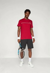 Raudona sportinė marškinė su baltu horizontaliu dryžiu, derinama su juodomis šortais. Modelis laiko teniso raketę, dėvi baltus sportbačius ir riešo juostas.