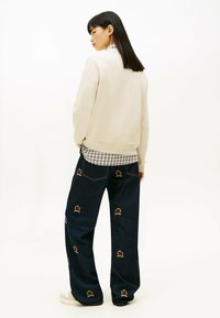 Felpa beige, jeans blu con ricamo di una corona colorata e sneakers bianche. Donna di spalle, camicia a quadri visibile sotto la felpa.