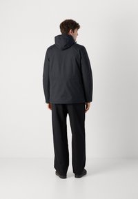 Veste noire à capuche avec une texture lisse, dotée d'une fermeture éclair et de coutures minimales, portée avec un pantalon noir à coupe ample et des chaussures sombres.