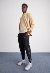 Beżowa koszula zapinana na guziki na jasnobeżowej koszulce, czarne spodnie cargo z bocznymi kieszeniami oraz szare sportowe buty z teksturowanym wzorem.