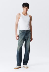 Young man standing wearing a white sleeveless shirt, faded blue jeans, and white sneakers against a plain light background.