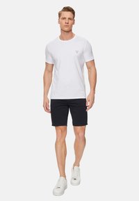 Camiseta de algodón blanca con un pequeño logotipo de triángulo rojo en el lado izquierdo del pecho, combinada con pantalones cortos negros y zapatillas blancas, en vista frontal.