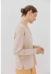 Beige langesleeve top met een V-hals, een losse pasvorm, zijsplitten aan de zoom en een zachte textuur, gecombineerd met een gele rok.