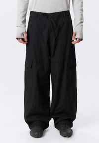 Personne portant un pantalon cargo noir ample avec des poches à rabat et des chaussures noires texturées, debout devant un fond blanc.