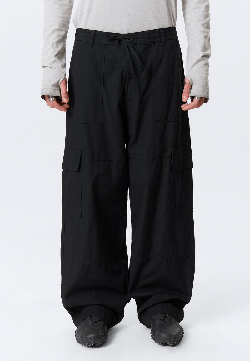 Personne portant un pantalon cargo noir ample avec des poches à rabat et des chaussures noires texturées, debout devant un fond blanc.