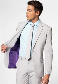 Homme portant un costume gris clair avec une cravate assortie et une chemise bleu clair, tenant la veste ouverte pour montrer la doublure violette à l’intérieur.