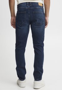 Vaqueros de mezclilla azul oscuro con corte recto, dos bolsillos traseros y una parche de cuero visible en la cintura. Combinados con zapatillas blancas.