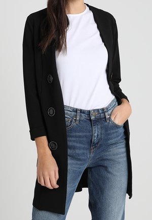 Vrouw draagt een zwarte jas met knopen over een wit t-shirt en blauwe jeans, met één hand in de zak en lang haar over de schouder.