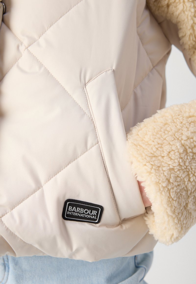 Giacca beige trapuntata con dettagli in fleece. Presenta un motivo diagonale, tasche laterali e un'etichetta del logo nera sul davanti. Polsini testurizzati.