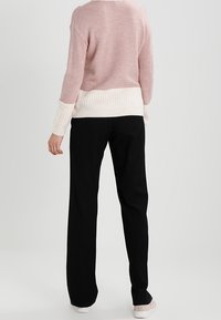 Person klädd i en rosa och vit stickad tröja, svarta vida byxor och ljusa skor, stående mot en enkel ljus bakgrund.