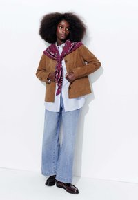 Veste en suédine marron à coupe décontractée, superposée à une chemise à rayures bleues et blanches. Associée à un jean large bleu clair et des chaussures foncées. Écharpe violette.