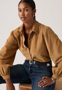 Brown corduroy button-up shirt with puff sleeves, paired with dark denim jeans and a black belt. Features silver jewelry accents.