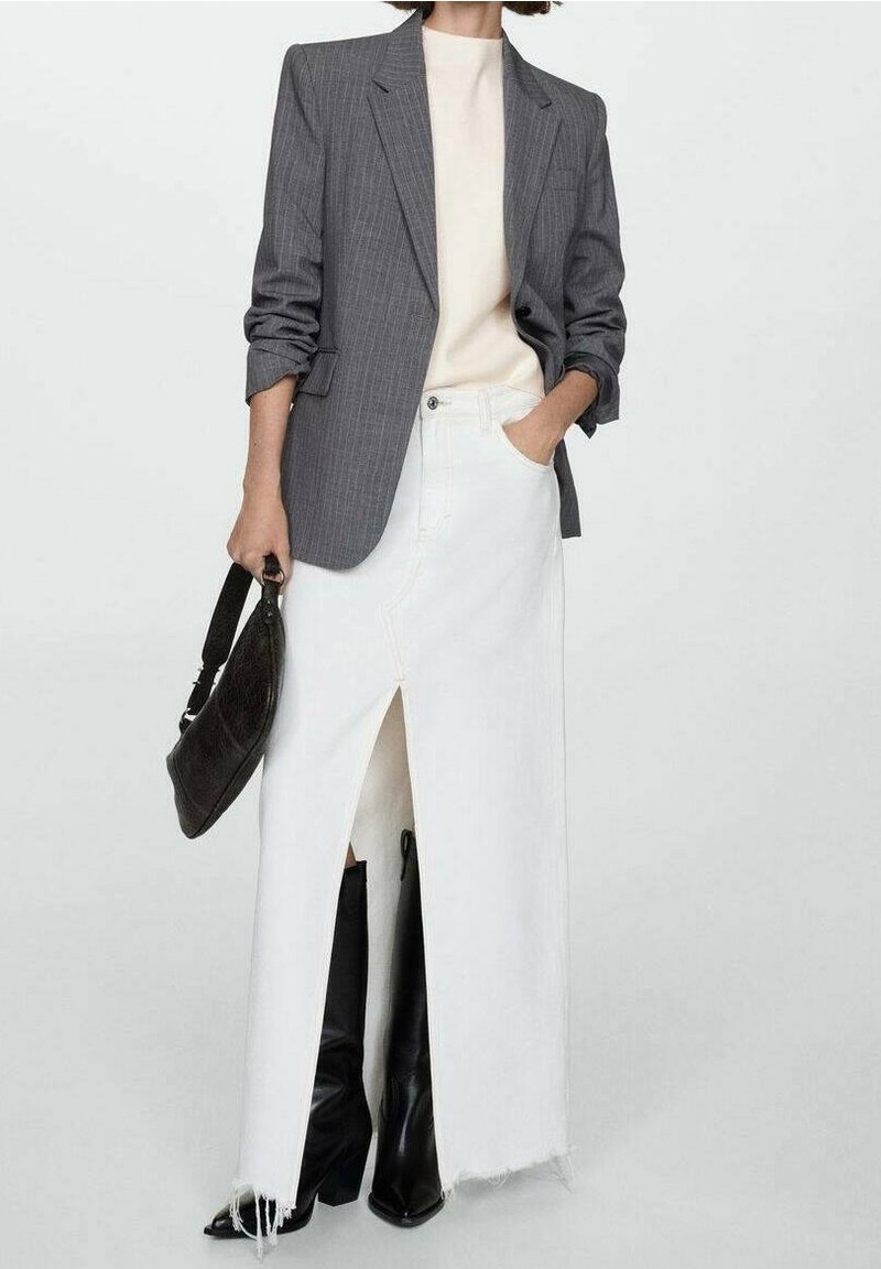 Femme portant un blazer gris à rayures fines sur un haut beige, une jupe blanche fendue haut, des bottes noires montant aux genoux, tenant un sac à main noir dans la main gauche.