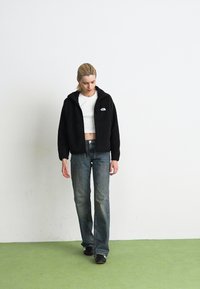 Young person walking indoors wearing a black fleece jacket, white crop top, loose jeans, and black sneakers on a green floor with a white wall background.