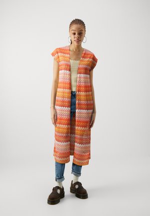 Cardigan kimono coloré, ouvert sur le devant, avec des motifs en zigzag orange, rose et blanc. Assorti à un haut en tricot et à un jean en denim.