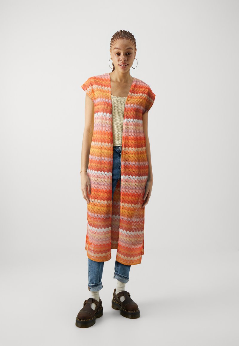 Cardigan kimono coloré, ouvert sur le devant, avec des motifs en zigzag orange, rose et blanc. Assorti à un haut en tricot et à un jean en denim.